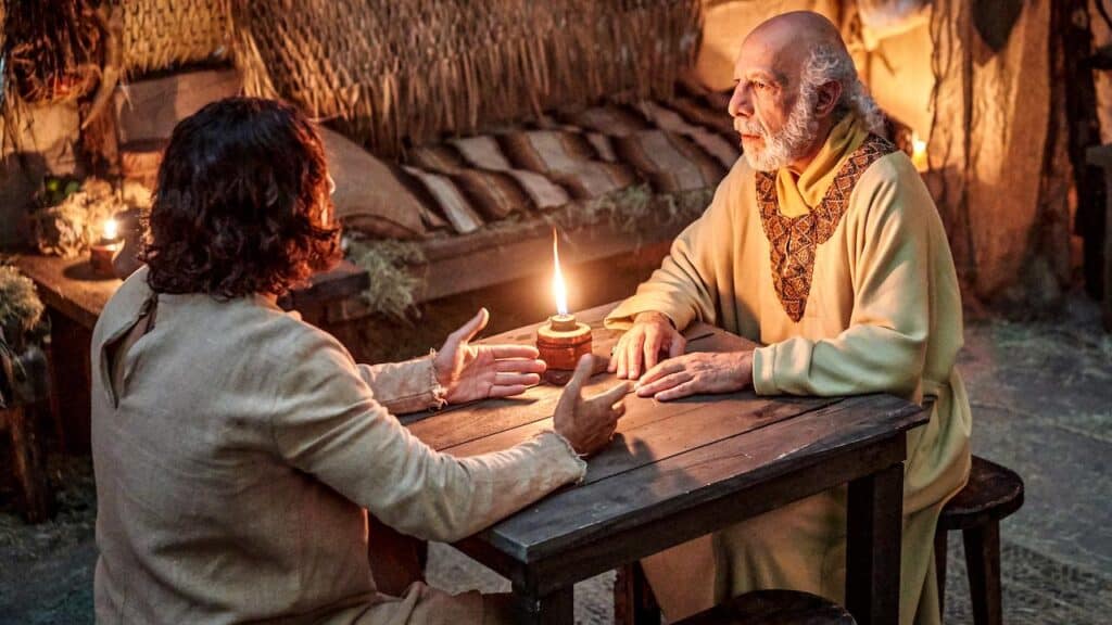 Two men in historical clothing sit across from each other at a wooden table, talking by candlelight in a rustic room—an intimate scene reminiscent of The Chosen that deepens faith through heartfelt conversation.