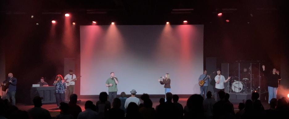 A band performs on stage with various instruments and microphones as an audience watches in a dimly lit venue with colored lights, while Christian worship song lyrics are examined for deeper meaning.