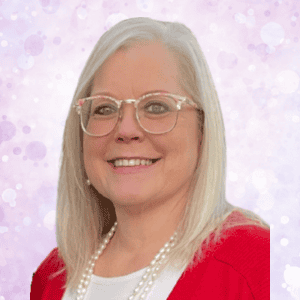 A smiling woman with straight, light blonde hair and glasses, wearing a red sweater, white blouse, and pearl necklace, stands in front of a light purple bokeh background.