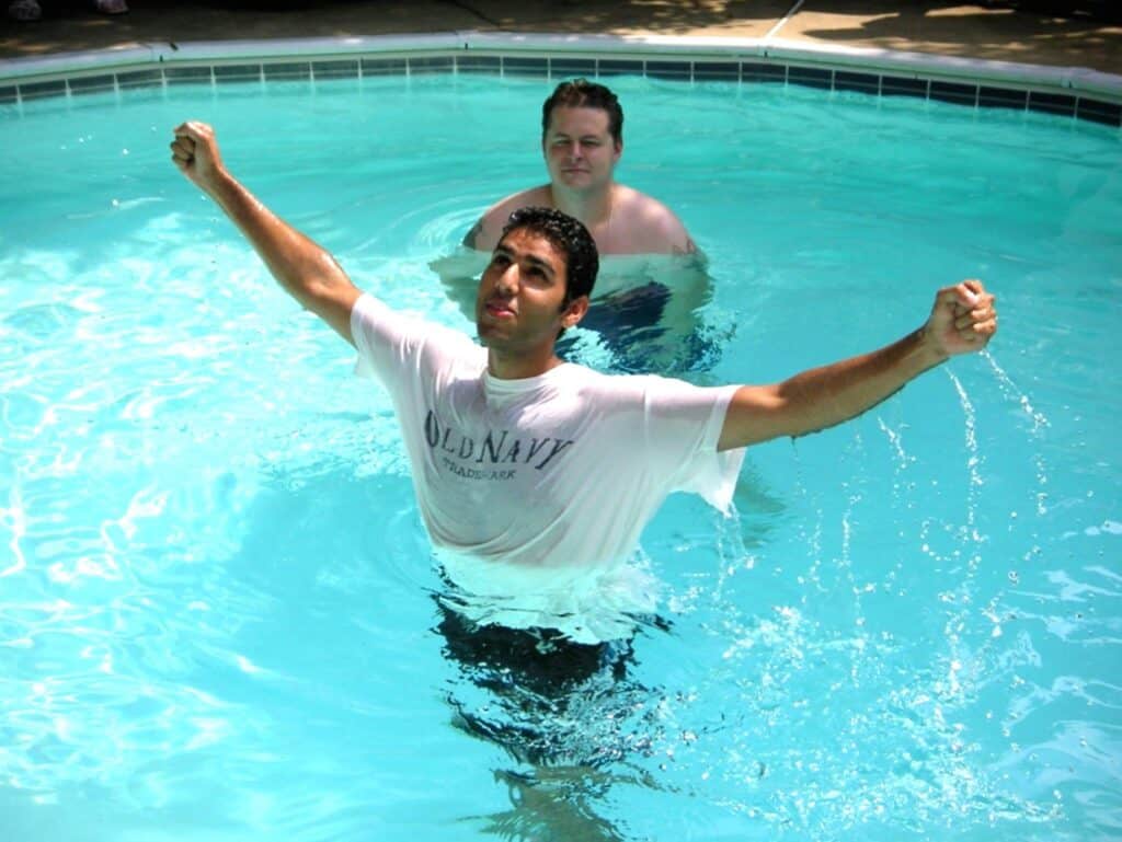 A man in a wet white T-shirt stands in a swimming pool with his arms raised and fists clenched, while another man watches behind him—capturing a triumphant moment reminiscent of themes from Nabeel Qureshi's Seeking Allah, Finding Jesus. Sunlight reflects off the pool’s surface.